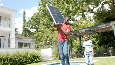 African-American-man-holds-a-solar-panel,-with-a-boy-looking-on-in-a-sunny-backyard-at-home
