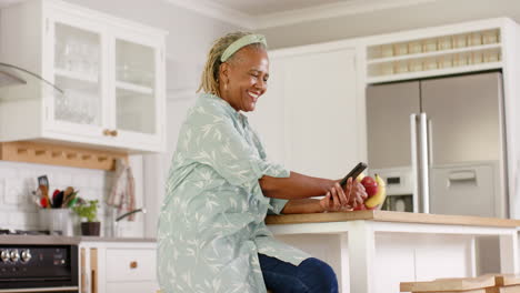 African-American-senior-woman-sitting-in-kitchen,-looking-at-smartphone
