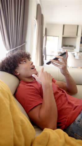 A-young-biracial-man-is-reclining-on-a-sofa,-interacting-with-his-smartphone