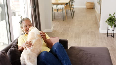 African-American-senior-woman-sitting-on-sofa,-petting-dog,-laughing