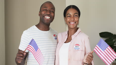 A-diverse-couple-is-beaming-with-pride,-holding-American-flags-and-wearing-vote-stickers