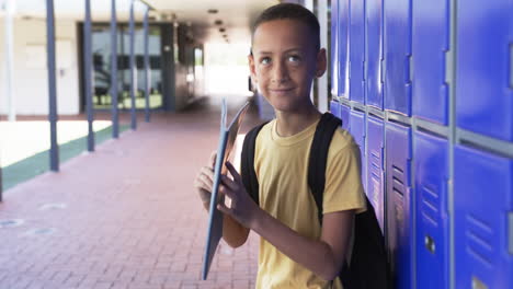 Biracial-boy-with-a-backpack-stands-by-blue-lockers,-holding-a-folder,-in-school
