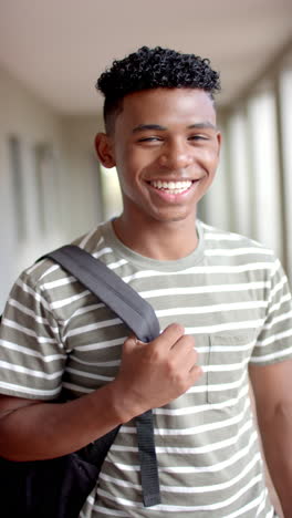 Vertical-video:-In-highschool,-biracial-male-teenager-with-a-bright-smile-holding-a-backpack-strap