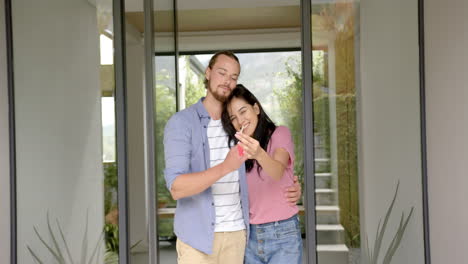 Diverse-couple-standing-at-home,-showing-keys-to-new-house