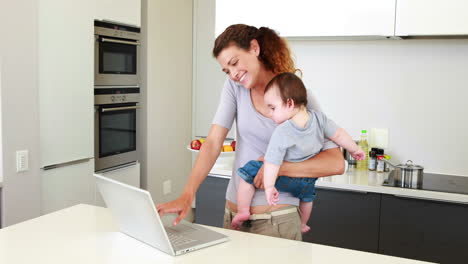 Mother-holding-her-happy-baby-boy-and-talking-on-phone-using-laptop