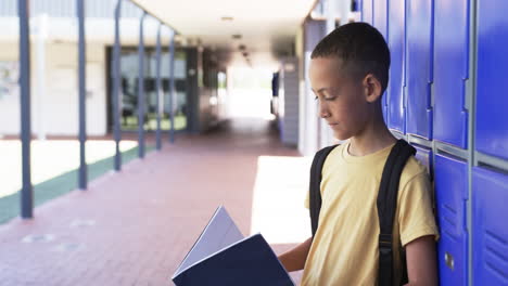 Biracial-boy-with-backpack-stands-by-blue-lockers,-reading-a-book,-copy-space