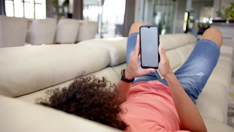 A-young-biracial-person-is-lying-on-sofa,-holding-a-smartphone