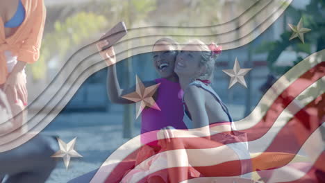 Animation-of-american-flag-elements-over-happy-diverse-female-friends-taking-selfies-on-sunny-beach