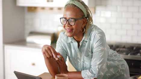 Afroamerikanische-Seniorin-Lehnt-An-Küchentheke-Und-Schaut-Auf-Tablet