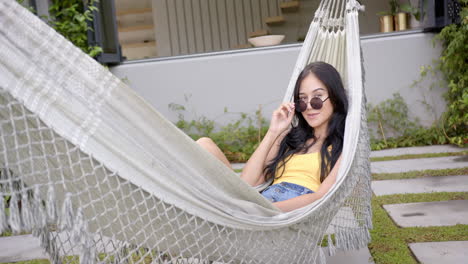 Una-Joven-Birracial-Descansa-En-Una-Hamaca-En-El-Patio-Trasero-De-Su-Casa,-Ajustando-Sus-Gafas-De-Sol