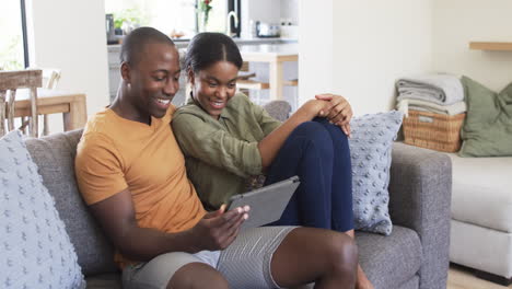 Ein-Junges-Afroamerikanisches-Paar-Sitzt-Zu-Hause-Auf-Der-Couch-Und-Lächelt,-Während-Es-Ein-Tablet-Verwendet