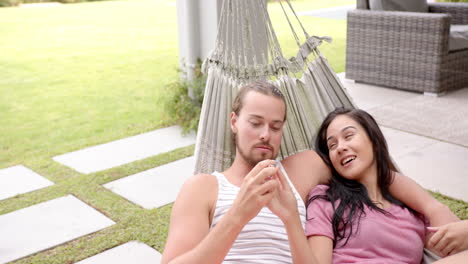 Diverse-couple-relaxing-in-hammock-outside-in-garden,-man-showing-smartphone,-copy-space