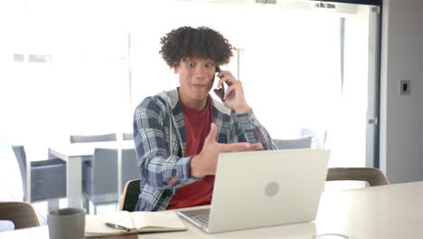 A-young-biracial-man-is-using-a-laptop-and-talking-on-the-phone-at-home-in-the-kitchen