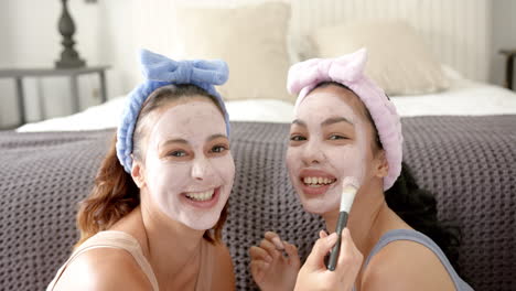 Two-young-biracial-female-friends-enjoy-a-skincare-routine,-wearing-facial-masks-and-headbands