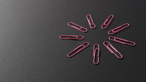 Pink-paper-clips-are-arranged-in-a-circular-pattern-on-a-black-background,-with-copy-space