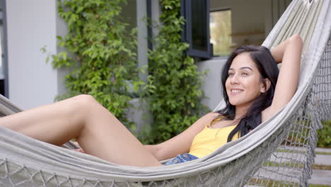 A-young-biracial-woman-relaxes-in-a-hammock-in-the-backyard-at-home,-copy-space