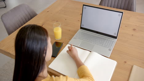 Asiatische-Biracial-Frau-Am-Tisch-Schreibt,-Mit-Laptop-Und-Smartphone-In-Der-Nähe,-Kopierraum