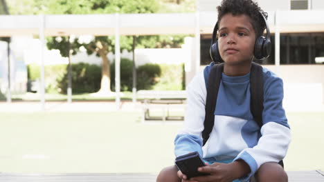 En-El-Patio-De-Una-Escuela,-Un-Joven-Estudiante-Afroamericano-Se-Sienta-Con-Auriculares,-Con-Espacio-Para-Copiar