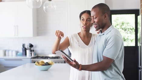 A-diverse-couple-discusses-in-their-kitchen,-using-tablet