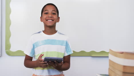 Biracial-boy-with-a-striped-shirt-holds-a-tablet-in-school,-smiling-in-a-classroom