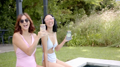 Two-biracial-female-friends-holding-drinks-by-pool-at-home,-copy-space
