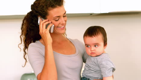 Mother-holding-her-baby-son-in-the-kitchen-talking-on-phone