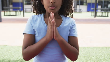 Biracial-girl-with-curly-hair-is-in-a-praying-pose-outdoors-at-school
