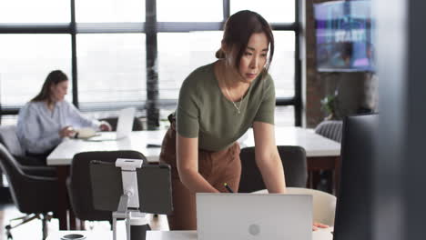 Asiatischer-Älterer-Arbeitet-Am-Laptop,-Junger-Kaukasischer-Kollege-Im-Hintergrund