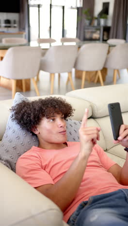 Vertical-video:-A-young-biracial-man-is-lounging-on-a-sofa,-smartphone-in-hand