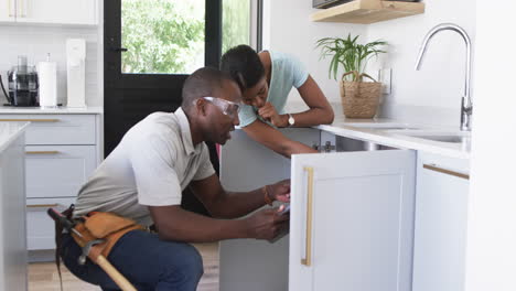 African-american-plumber-repairing-kitchen-sink-in-home-of-woman