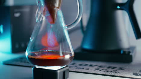 Pour-Over-Methode---Kaffee-Tropft-Aus-Dem-Filterpapier-In-Die-Chemex-Kaffeemaschine,-Nahaufnahme,-Im-Hintergrund-Ein-Barista-Wasserkessel