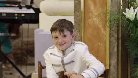 Boy-In-A-White-Ceremonial-Suit-Sitting-And-Smiling-During-Communion-Celebration