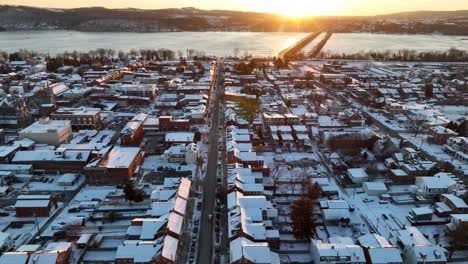Aerial-view-of-a-snowy-town-at-sunrise,-with-a-frozen-river-and-golden-sunlight-reflecting-off-the-icy-surface