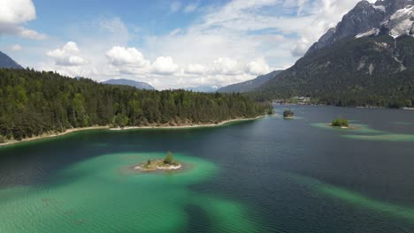 Aerial-view-of-the-colorful-Eibsee-Lake-in-Bayern,-Germany,-surrounded-by-pine-trees-and-a-distant-mountain-range,-highlighting-the-tranquil-harmony-of-nature