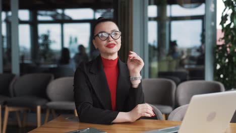 beautiful-woman-in-business-attire-puts-on-eyeglasses-while-sitting-in-a-restaurant-with-a-laptop