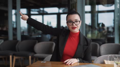 stern-young-woman-in-business-attire-angrily-gestures-with-her-hands-and-argues,-looking-into-the-camera-while-sitting-in-a-stylish-restaurant