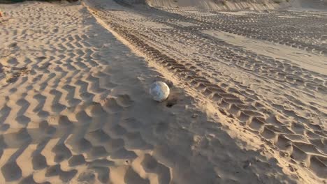 Una-Vieja-Pelota-De-Fútbol-Desaliñada-Rodando-Por-La-Arena-De-La-Playa-Con-Huellas-De-Neumáticos-En-La-Arena-En-Un-Día-Soleado