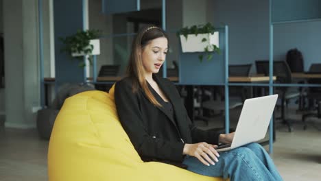 lovely-young-woman-in-business-attire-sits-with-her-laptop-in-a-stylish,-modern-coworking-office,-greets-via-video-call-through-the-laptop,-and-starts-a-conversation