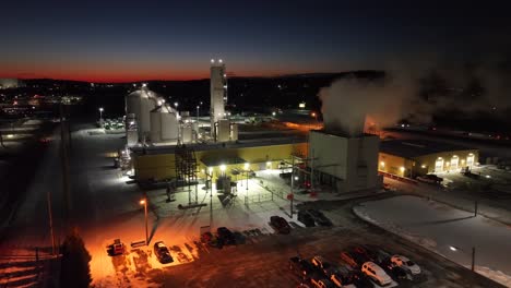 Aerial-view-of-an-industrial-facility-at-night-with-steam-and-lights,-under-a-colorful-sunset-sky-in-the-USA