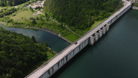 Black-car-driving-on-a-dam,-between-a-water-reservoir-and-deep-valley,-tracking-drone-wide-shot