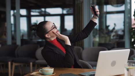 Una-Hermosa-Mujer-Vestida-De-Negocios-Está-Haciendo-Una-Videollamada-En-Su-Teléfono-Inteligente-Con-Una-Sonrisa-Mientras-Está-Sentada-Frente-A-Su-Computadora-Portátil