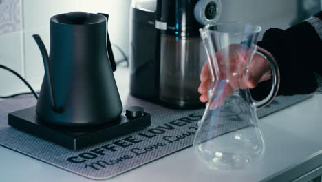 Legen-Sie-Einen-Papierfilter-In-Eine-Chemex-Kaffeemaschine-Aus-Glas,-Bevor-Sie-Einen-Filterkaffee-Zubereiten,-Im-Hintergrund-Ein-Barista-Wasserkocher-Und-Eine-Kaffeemühle
