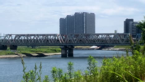 Der-Zug-überquert-An-Einem-Sonnigen-Tag-Eine-Stahlbrücke-über-Einen-Ruhigen-Fluss-Mit-Hohen-Gebäuden-Im-Hintergrund