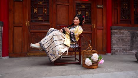 Eine-Frau-Aus-Der-Qing-Dynastie-Lächelt,-Während-Sie-Mit-Ihrem-Haar-Spielt,-Trägt-Traditionelle-Kleidung-Und-Sitzt-Vor-Einer-Verzierten-Holztür-Mit-Einem-Vogelkäfig-Und-Blumen-In-Der-Nähe-Elegant-Auf-Einem-Stuhl