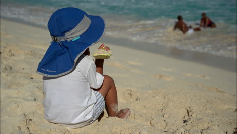 Nicht-Erkennbarer-Mexikanischer-Lateinamerikanischer-Junge-Mit-Hut-Und-Weißem-T-Shirt,-Der-Auf-Dem-Sand-Sitzt-Und-Mit-Einer-Plastikschaufel-Spielt-Und-Das-Meer-Beobachtet