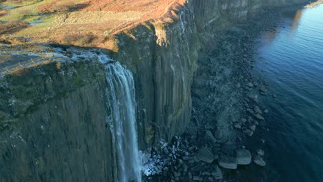 Waterfall-in-shadow-cascading-down-steep-cliff-to-frosty-and-stony-beach,-early-morning-in-winter-with-sunlit-moorland-top-at-Kilt-Rock-Waterfall,-Isle-of-Skye,-Western-Highlands,-Scotland,-UK