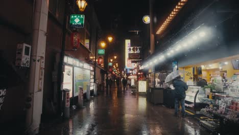 Lluvia-Cayendo-Sobre-El-Mercado-Callejero-Durante-La-Noche-Con-Gente-Sosteniendo-Paraguas-En-Osaka,-Japón