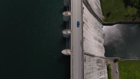 Convertible-blue-car-driving-on-a-dam,-between-a-water-reservoir-and-deep-valley,-tracking-drone-shot