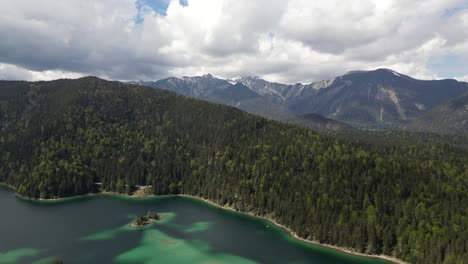 Aerial-view-of-the-vibrant-Eibsee-Lake-in-Bayern,-Germany,-surrounded-by-natural-pine-forests-and-a-distant-mountain-range,-embodying-the-serene-harmony-of-nature