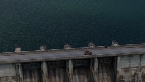 Orange-suv-driving-on-a-dam,-between-a-water-reservoir-and-deep-valley,-tracking-drone-shot